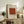 May include: Framed beige poster with rust-colored geometric pattern grid displayed on weathered wood sideboard. Warm neutral living room setting with cream upholstered chair, brown velvet pillows, potted fern, ceramic vase, and stacked books. Natural lighting creates soft shadows on white wall.
