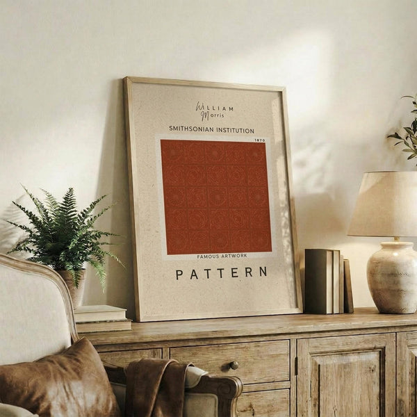 May include: Framed beige poster with rust-colored geometric pattern grid displayed on weathered wood sideboard. Warm neutral living room setting with cream upholstered chair, brown velvet pillows, potted fern, ceramic vase, and stacked books. Natural lighting creates soft shadows on white wall.