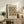 May include: Framed William Morris textile pattern print in earth tones displayed on rustic wooden sideboard alongside fern plant, ceramic vase, lamp, and books in cozy living room setting with beige armchair and warm natural lighting