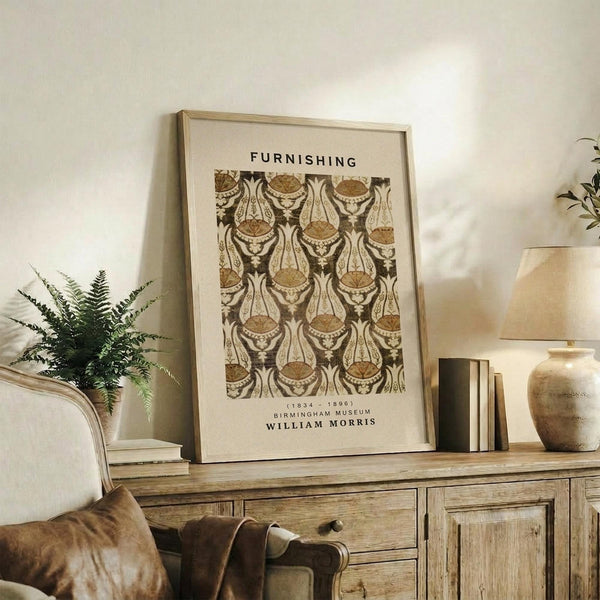 May include: Framed William Morris textile pattern print in earth tones displayed on rustic wooden sideboard alongside fern plant, ceramic vase, lamp, and books in cozy living room setting with beige armchair and warm natural lighting