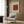 May include: Beige framed poster with rust-colored geometric pattern square mounted on wood paneled wall above cream upholstered armchair with geometric pillow. Modern reading corner features brass floor lamp and dark round side table with open book near window with flowing curtains.