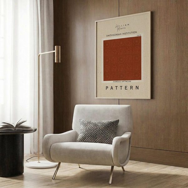May include: Beige framed poster with rust-colored geometric pattern square mounted on wood paneled wall above cream upholstered armchair with geometric pillow. Modern reading corner features brass floor lamp and dark round side table with open book near window with flowing curtains.