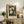 May include: Framed botanical poster with cream-colored mat displaying white and pale flowers with green foliage on dark background. Light wood frame sits on rustic wooden sideboard next to fern plant, books, ceramic vase and table lamp in warm neutral-toned living room setting.