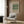 May include: Beige framed poster with green botanical William Morris tulip pattern displayed on wood-paneled wall above cream upholstered armchair with geometric pillow. Modern reading nook with brass floor lamp, dark round side table with open book, and white curtains. Warm neutral interior styling.