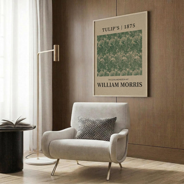 May include: Beige framed poster with green botanical William Morris tulip pattern displayed on wood-paneled wall above cream upholstered armchair with geometric pillow. Modern reading nook with brass floor lamp, dark round side table with open book, and white curtains. Warm neutral interior styling.