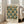May include: Framed poster with ornate Victorian wallpaper pattern in teal, cream and gold tones featuring repeated decorative motifs. White wooden frame leans against bright living room wall next to beige upholstered sofa. Natural sunlight streams through large windows creating soft shadows on hardwood floors.