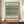 May include: Framed vintage botanical poster with green leafy tulip pattern on cream background, displayed on wooden sideboard next to white ceramic vase and stacked books against beige paneled wall in elegant room setting.