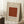 May include: Framed vintage-style poster with beige background featuring William Morris, Smithsonian Institution text, and rust-colored square displaying intricate circular floral pattern. Frame leans against white wall beside wooden side table with white coffee cup and saucer. Cream textured rug and beige fabric visible in cozy interior setting.