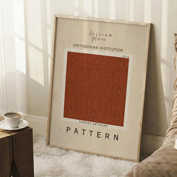 May include: Framed vintage-style poster with beige background featuring William Morris, Smithsonian Institution text, and rust-colored square displaying intricate circular floral pattern. Frame leans against white wall beside wooden side table with white coffee cup and saucer. Cream textured rug and beige fabric visible in cozy interior setting.