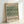 May include: Framed poster print featuring William Morris's Tulip pattern from 1875 in muted green and cream tones. The vintage botanical wallpaper design shows repeating tulip and foliage motifs. Print displays "TULIP'S | 1875" at top and "WILLIAM MORRIS" with "THE CLEVELAND MUSEUM ART" text below the pattern. Light wood frame rests against white wall on pale surface, creating soft natural shadows.