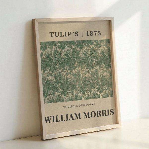 May include: Framed poster print featuring William Morris's Tulip pattern from 1875 in muted green and cream tones. The vintage botanical wallpaper design shows repeating tulip and foliage motifs. Print displays "TULIP'S | 1875" at top and "WILLIAM MORRIS" with "THE CLEVELAND MUSEUM ART" text below the pattern. Light wood frame rests against white wall on pale surface, creating soft natural shadows.