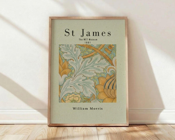 May include: Framed poster print in natural wood frame leaning against white wall on light wooden floor. Features botanical design with ornate sage green leaves and golden orange decorative elements in William Morris style. Text reads "St James The MET Museum 1881" at top and "William Morris" at bottom on pale green background. Soft natural lighting creates diagonal shadows across white textured wall.
