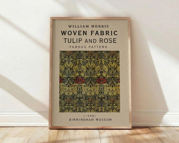 May include: Framed vintage botanical textile design poster leaning against white wall on light wood floor. The artwork features intricate Arts and Crafts pattern with stylized tulips and roses in deep greens, golden yellows, and burgundy red on cream background. Text reads "William Morris Woven Fabric Tulip and Rose Famous Pattern (1890) Birmingham Museum" in black typography above and below the decorative fabric sample.