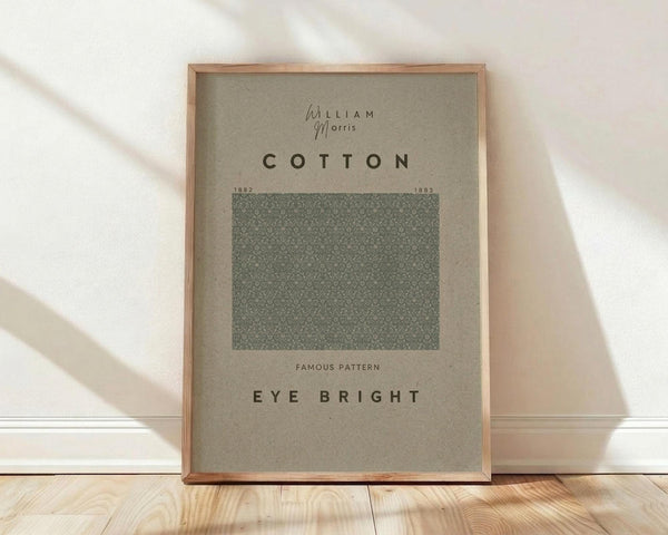 May include: Framed poster with beige background displaying "William Morris" and "COTTON" text above a green geometric pattern sample, labeled "EYE BRIGHT" below. Natural wood frame sits on light wooden floor against white wall with soft sunlight shadows.