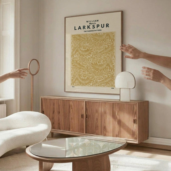 May include: Framed vintage botanical poster with golden yellow leaf pattern on cream background, displayed above mid-century wooden credenza in bright living room. Hands positioning artwork on white wall, with modern white chair, glass coffee table, and contemporary lamp creating warm, minimalist interior styling.