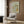 May include: Beige armchair with geometric patterned pillow positioned next to gold floor lamp and dark side table with open book. Framed vintage botanical art print featuring green and orange flowing leaf patterns hangs on wood-paneled wall. Modern minimalist living room setting with neutral tones and natural lighting from nearby window with curtains.