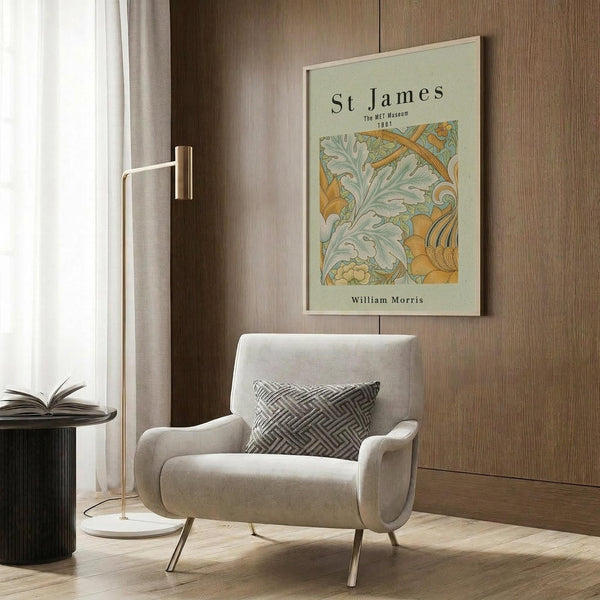 May include: Beige armchair with geometric patterned pillow positioned next to gold floor lamp and dark side table with open book. Framed vintage botanical art print featuring green and orange flowing leaf patterns hangs on wood-paneled wall. Modern minimalist living room setting with neutral tones and natural lighting from nearby window with curtains.