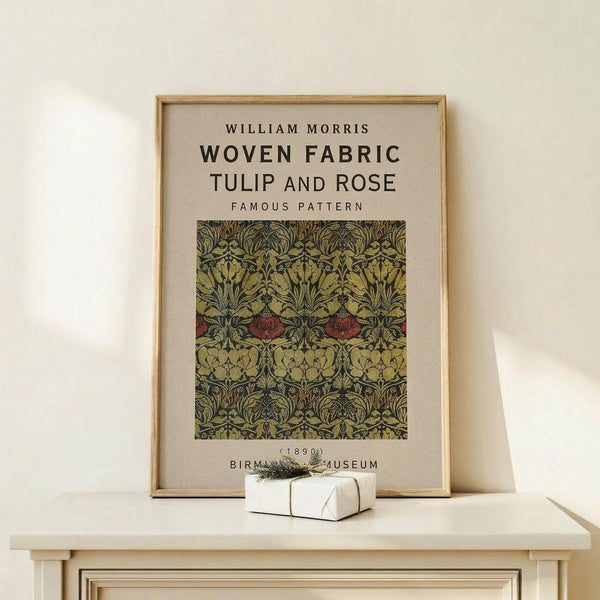 May include: Framed vintage botanical poster featuring William Morris's intricate tulip and rose pattern in olive green, burgundy red, and gold tones, displayed on a light wood console table beside a small white wrapped gift with evergreen sprig against a cream wall.