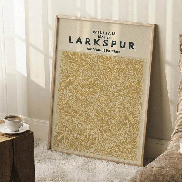 May include: Framed botanical art print in golden yellow and cream featuring William Morris Larkspur pattern with intricate flowing leaves and flowers. Light wood frame leaning against white paneled wall next to wooden stool with white coffee cup, beige throw blanket visible in corner on light textured rug.