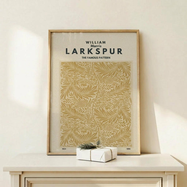 May include: Framed botanical print with golden yellow intricate leaf and fern pattern against cream background, displayed on light wood console table next to small white wrapped gift with evergreen sprig. The artwork features flowing organic motifs in warm tones within a natural wood frame.