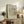 May include: Framed museum-style poster featuring William Morris botanical artwork with flowing green acanthus leaves and golden yellow flowers on pale mint background. Light wood frame displayed on weathered wooden sideboard alongside potted fern, ceramic vase, and stacked books in warm, natural living room setting.