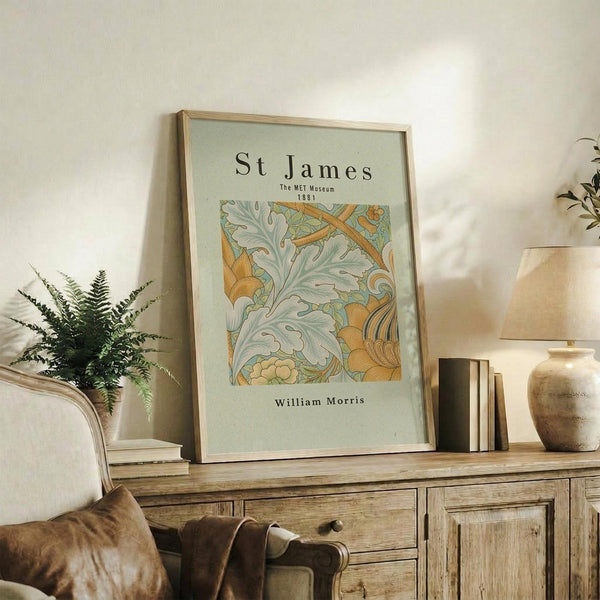 May include: Framed museum-style poster featuring William Morris botanical artwork with flowing green acanthus leaves and golden yellow flowers on pale mint background. Light wood frame displayed on weathered wooden sideboard alongside potted fern, ceramic vase, and stacked books in warm, natural living room setting.