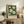 May include: Framed botanical poster with white magnolia flowers on dark green background, displayed on rustic wooden sideboard alongside fern plant, ceramic vase, and books in warm-toned living room setting with beige chair and neutral walls