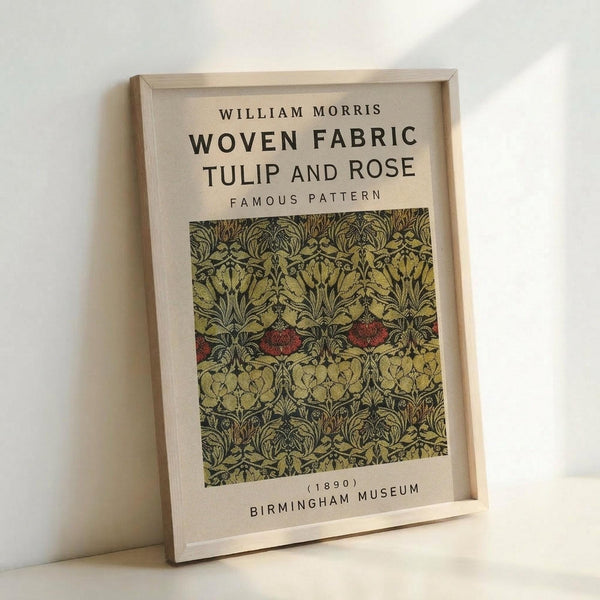 May include: Framed poster print leaning against white wall showing William Morris woven fabric pattern from 1890. Features intricate botanical design with yellow tulips, red roses, and green foliage on cream background. Light wood frame with museum-style labeling. Soft natural lighting creates gentle shadows on textured wall surface.