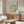 May include: Framed William Morris botanical poster with sage green background and ornate floral pattern in orange and teal tones, displayed above mid-century walnut credenza with sliding doors. Modern white sculptural chair and oval glass coffee table visible in minimalist living room with cream walls and natural lighting.