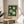 May include: Framed botanical poster featuring white magnolia flowers with green leaves on dark emerald background, displayed on round wooden side table next to beige upholstered chair. Natural lighting creates soft shadows across white walls. Plant leaves visible in corner of minimalist room setting.