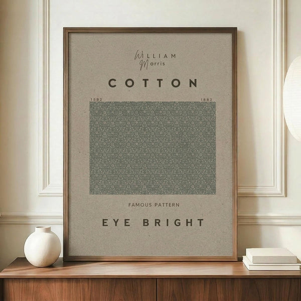 May include: Framed botanical poster with green Eye Bright pattern sample displayed on wooden credenza. Features William Morris branding, cotton fabric dates 1882-1883, and "Famous Pattern" text on beige background. Styled with white ceramic vase and books against cream paneled wall.