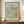 May include: Framed botanical art print featuring William Morris's St James pattern from 1881, displayed on wooden sideboard beside white ceramic vase and stacked books. The artwork shows flowing acanthus leaves in sage green and golden orange tones against cream background, housed in thin brown frame against white wainscoted wall.