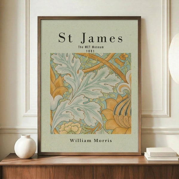 May include: Framed botanical art print featuring William Morris's St James pattern from 1881, displayed on wooden sideboard beside white ceramic vase and stacked books. The artwork shows flowing acanthus leaves in sage green and golden orange tones against cream background, housed in thin brown frame against white wainscoted wall.