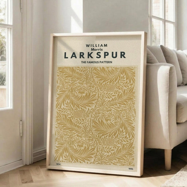 May include: Large framed poster with William Morris Larkspur pattern in golden yellow and cream, featuring intricate botanical design with flowing leaves and flowers. Light wood frame leans against white wall corner next to beige upholstered chair with wooden legs on light hardwood floor in bright, airy room with tall windows.