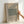 May include: Framed poster with beige background displaying "William Morris" and "COTTON" text above a sage green textured fabric pattern sample, dated 1882, with "FAMOUS PATTERN EYE BRIGHT" below. Light wood frame held by hand against white wall in natural lighting.