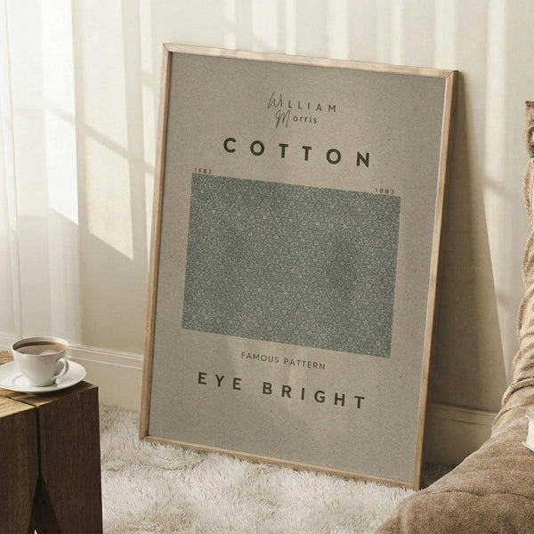 May include: Framed botanical poster leaning against white wall featuring green textured pattern sample labeled "Cotton" and "Eye Bright" with William Morris attribution and dates 1882-1883. Displayed beside wooden block with white coffee cup and saucer, neutral textured rug, and beige throw blanket in soft natural lighting.