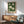 May include: Framed botanical poster with white magnolia flowers on deep green background displayed above wooden sideboard in modern living room. Mid-century style armchair in sage green fabric with wooden arms sits nearby. Natural sunlight creates shadows on white wall. Minimalist decor with table lamp and round coffee table visible.