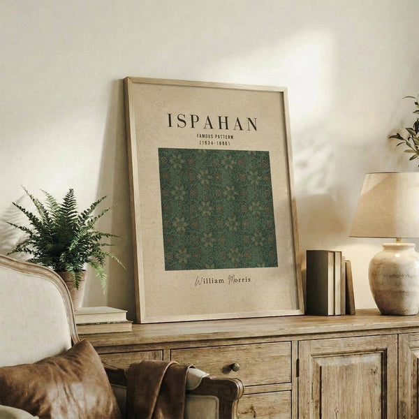 May include: Framed poster with beige background displaying "ISPAHAN" text and green floral pattern square, attributed to William Morris with dates 1834-1888, leaning against white wall on rustic wooden sideboard alongside fern plant, books, ceramic vase, and brown leather chair visible in cozy living room setting with warm natural lighting.