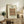 May include: Framed art print displaying ornate peacock and dragon pattern in muted pink, green, and blue tones, positioned on rustic wooden sideboard alongside potted fern, stacked books, ceramic vase, and table lamp in neutral-toned living room with beige upholstered furniture