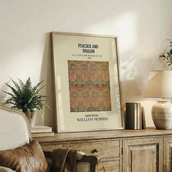 May include: Framed art print displaying ornate peacock and dragon pattern in muted pink, green, and blue tones, positioned on rustic wooden sideboard alongside potted fern, stacked books, ceramic vase, and table lamp in neutral-toned living room with beige upholstered furniture