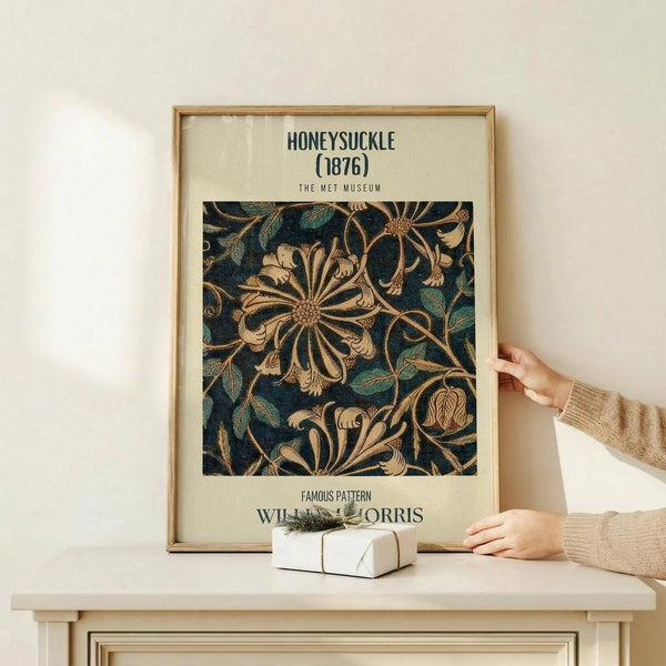 May include: Framed vintage botanical poster with intricate honeysuckle pattern in deep navy, teal and golden tones, displayed on light wood console table. Person in beige sweater holds corner of wooden frame. Small wrapped gift with twine and greenery sits on table surface against cream wall background.