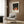 May include: Framed poster featuring spotted deer in stylized forest scene with dark starry sky and red dots, displayed above beige upholstered armchair with geometric patterned pillow. Modern living room setting with brass floor lamp, dark round side table with open book, wood panel wall, and light curtains. White matted frame against warm neutral interior.