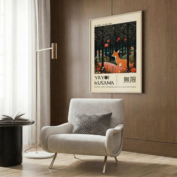 May include: Framed poster featuring spotted deer in stylized forest scene with dark starry sky and red dots, displayed above beige upholstered armchair with geometric patterned pillow. Modern living room setting with brass floor lamp, dark round side table with open book, wood panel wall, and light curtains. White matted frame against warm neutral interior.