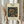May include: Hands holding a framed vintage William Morris floral pattern print featuring ornate honeysuckle flowers and leaves in gold, teal, and navy blue against cream matting. Light wood frame, dried wheat stems visible on left, soft natural lighting through white curtains creating gentle shadows on neutral background.