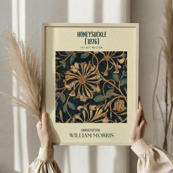 May include: Hands holding a framed vintage William Morris floral pattern print featuring ornate honeysuckle flowers and leaves in gold, teal, and navy blue against cream matting. Light wood frame, dried wheat stems visible on left, soft natural lighting through white curtains creating gentle shadows on neutral background.