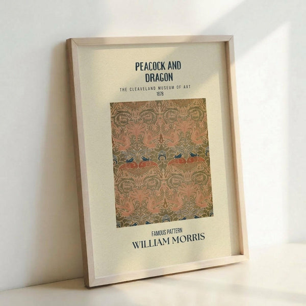 May include: Framed vintage-style poster featuring William Morris's ornate Peacock and Dragon textile pattern from 1878. The design shows intricate interwoven peacocks and mythical creatures in muted coral, sage green, and gold tones against cream background. Displayed in light wood frame leaning against white wall, showing museum-quality reproduction with classic typography identifying the Cleveland Museum of Art collection piece.