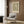 May include: Framed botanical poster with green and cream leaf pattern hanging on wood paneled wall above beige upholstered armchair with geometric patterned pillow, brass floor lamp, and dark round side table with open book in modern neutral-toned room setting