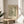 May include: Framed botanical art print featuring William Morris-style flowing foliage pattern in sage green and cream tones, displayed on light wood round side table next to beige upholstered chair. Soft natural lighting creates shadows across white walls, with houseplant visible in corner of minimalist room setting.