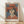 May include: Framed art print featuring a woman wearing an orange wide-brimmed hat and coral-colored top with white collar detail against a blue-gray background, painted in a modernist style reminiscent of early 20th century portraiture. The wooden frame sits against a white textured wall on light wooden flooring with natural sunlight casting diagonal shadows across the scene.