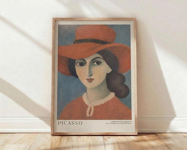 May include: Framed art print featuring a woman wearing an orange wide-brimmed hat and coral-colored top with white collar detail against a blue-gray background, painted in a modernist style reminiscent of early 20th century portraiture. The wooden frame sits against a white textured wall on light wooden flooring with natural sunlight casting diagonal shadows across the scene.